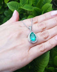 Hand holding a white gold necklace with a natural emerald pendant against a green leafy background.