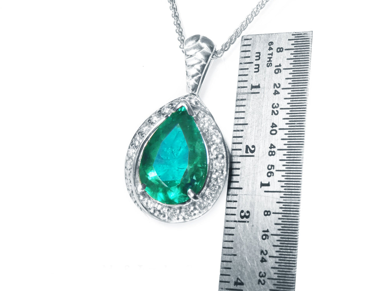 Pendant with pear cut emerald and white gold chain next to a ruler for size reference on a white background