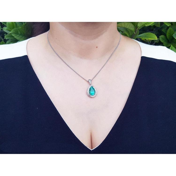 Pear-shaped natural emerald pendant with diamond halo on a 14K white gold chain, worn on a woman's neck.