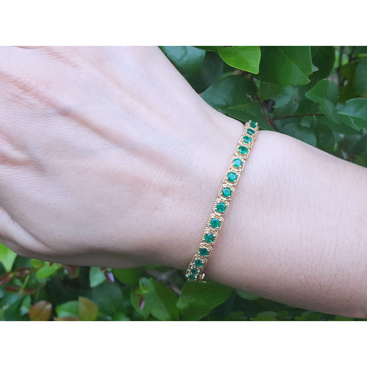 Gold bracelet with round cut natural colombian emeralds on a wrist against a green leafy background.