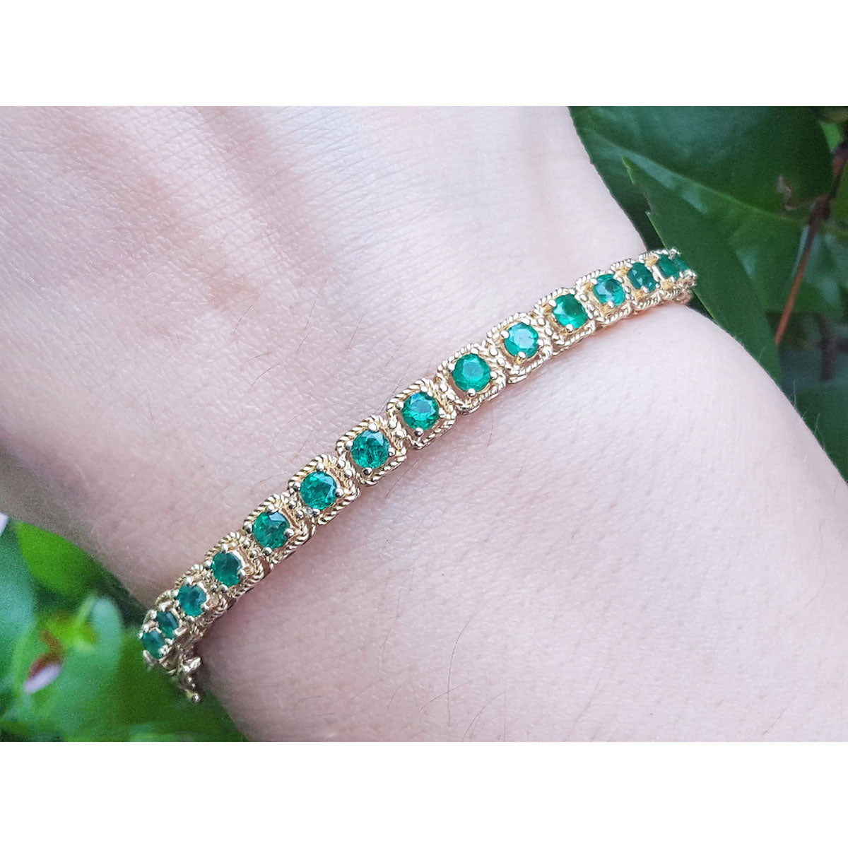 A round cut natural emeralds prong set bangle bracelet crafted in 14k yellow gold with rope decoration worn on a wrist.