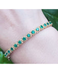 A round cut natural emeralds prong set bangle bracelet crafted in 14k yellow gold with rope decoration worn on a wrist.