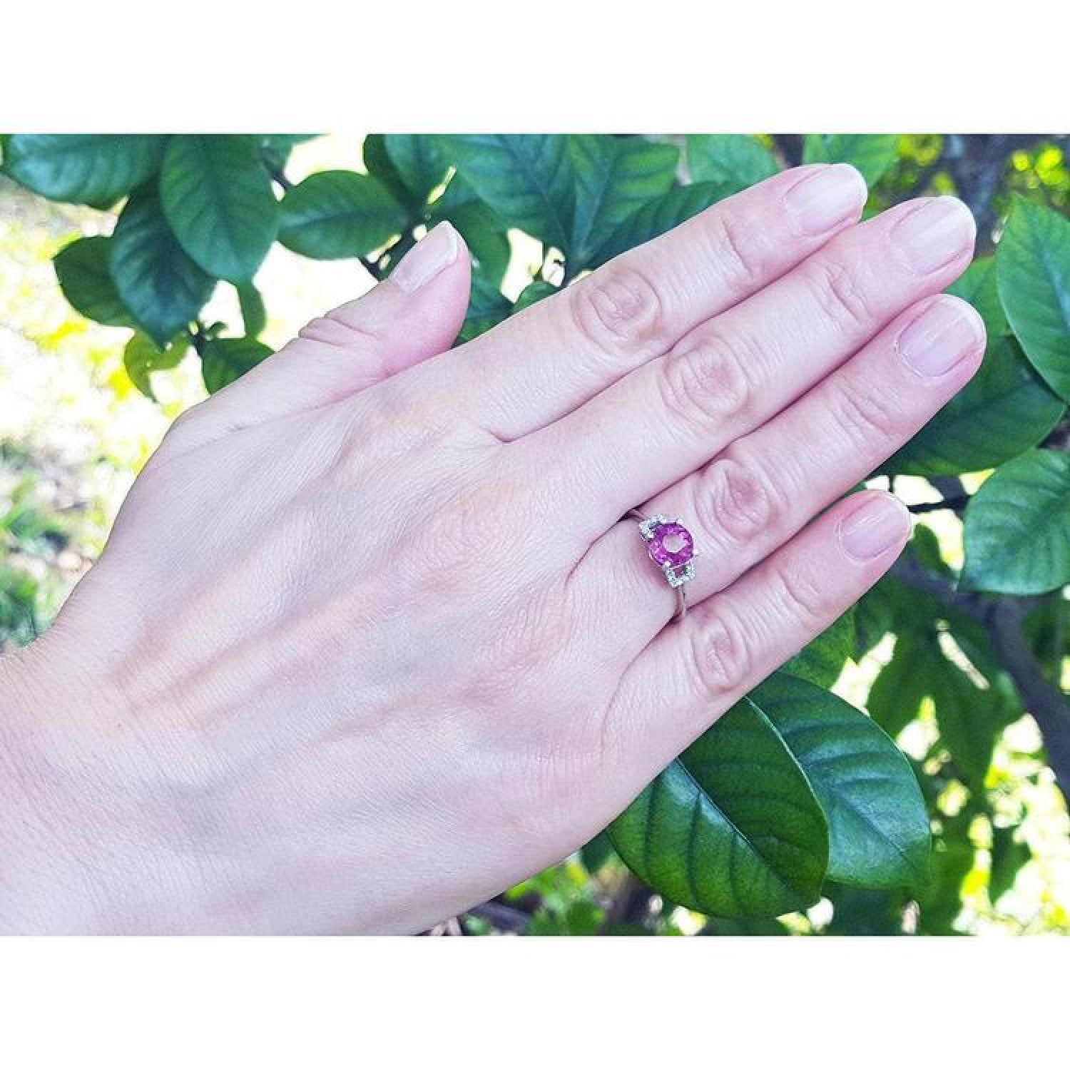 Round cut natural ruby ring with side diamonds in 14k white gold on a hand, surrounded by green leaves.
