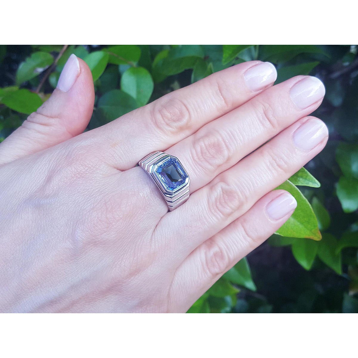 Hand wearing a white gold ring with a blue sapphire gemstone against a green leafy background