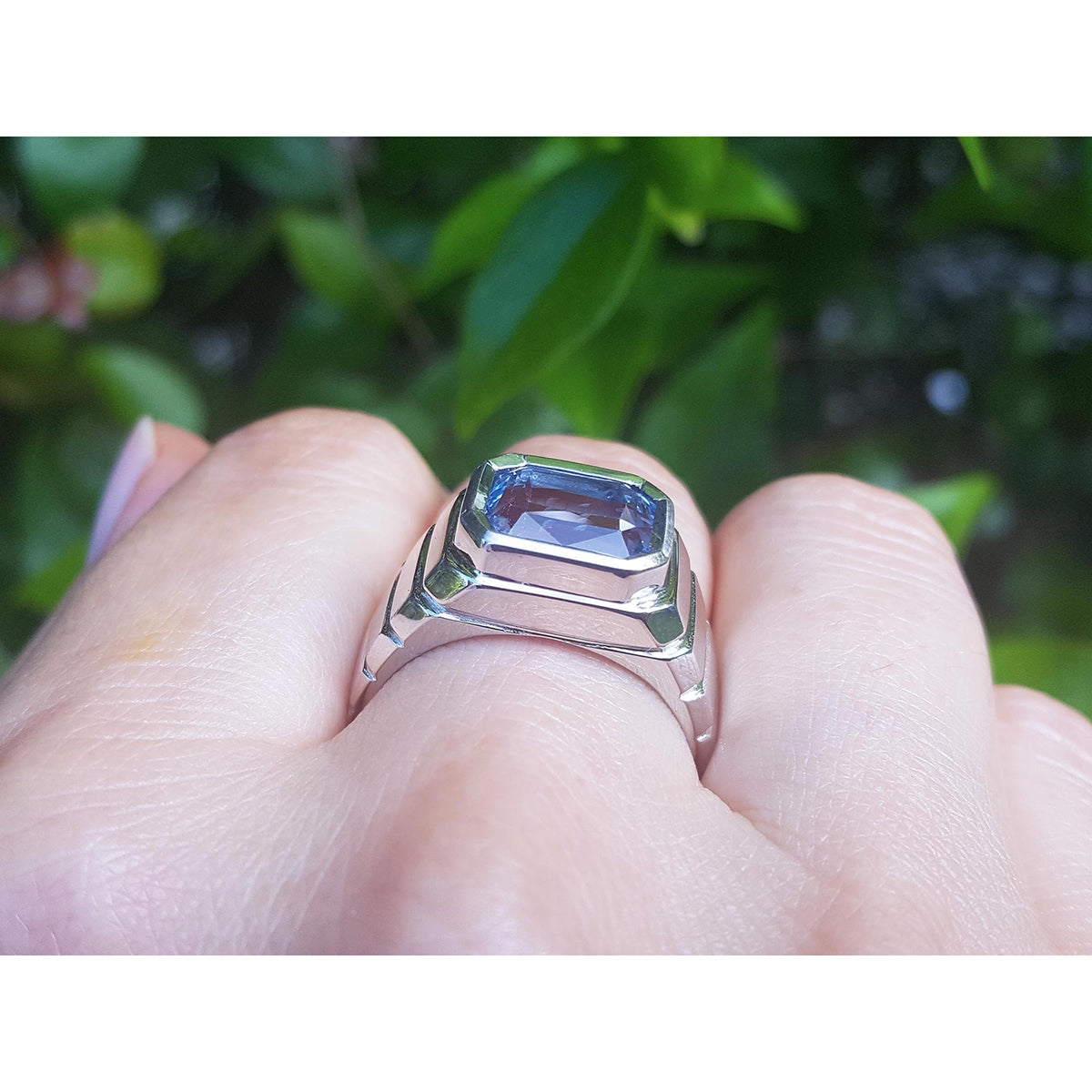 18k white gold ring with a blue natural sapphire worn on a finger against a green leafy background