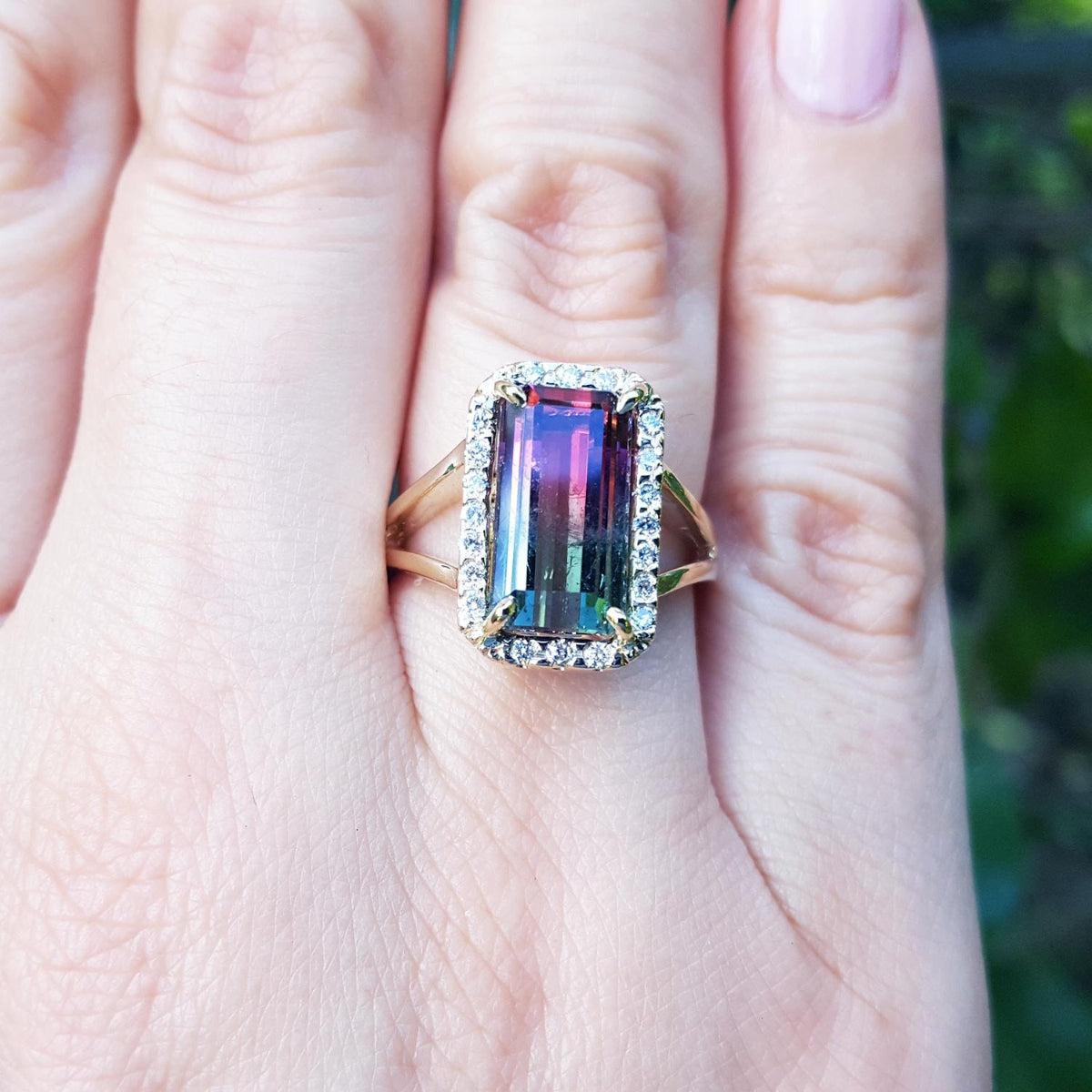 A hand wearing a natural watermelon tourmaline ring with a halo diamond, crafted in 14k yellow gold, on a green blurry background.