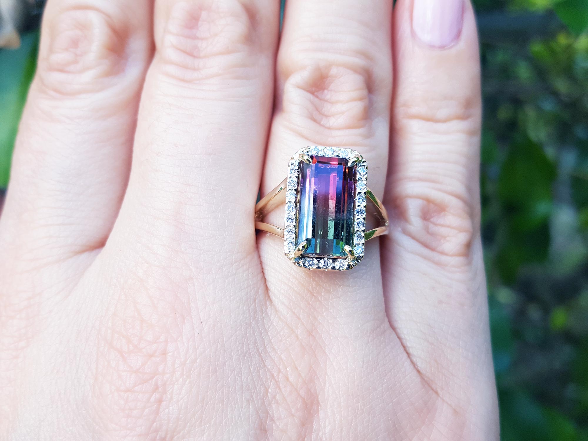 A hand wearing a natural watermelon tourmaline ring with a halo diamond, crafted in 14k yellow gold, on a green blurry background.