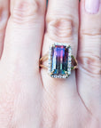 A hand wearing a natural watermelon tourmaline ring with a halo diamond, crafted in 14k yellow gold, on a green blurry background.