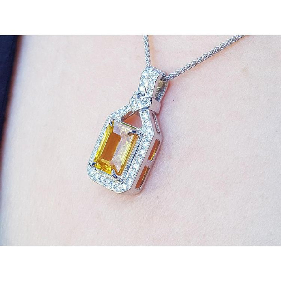 Emerald cut yellow sapphire pendant with halo diamonds on an 18K white gold chain, worn on a woman's neck.