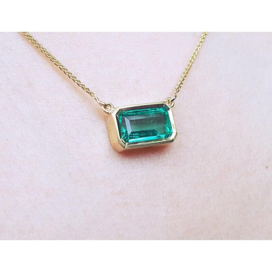 East-west emerald-cut solitaire emerald necklace in 18K yellow gold, bezel set.
