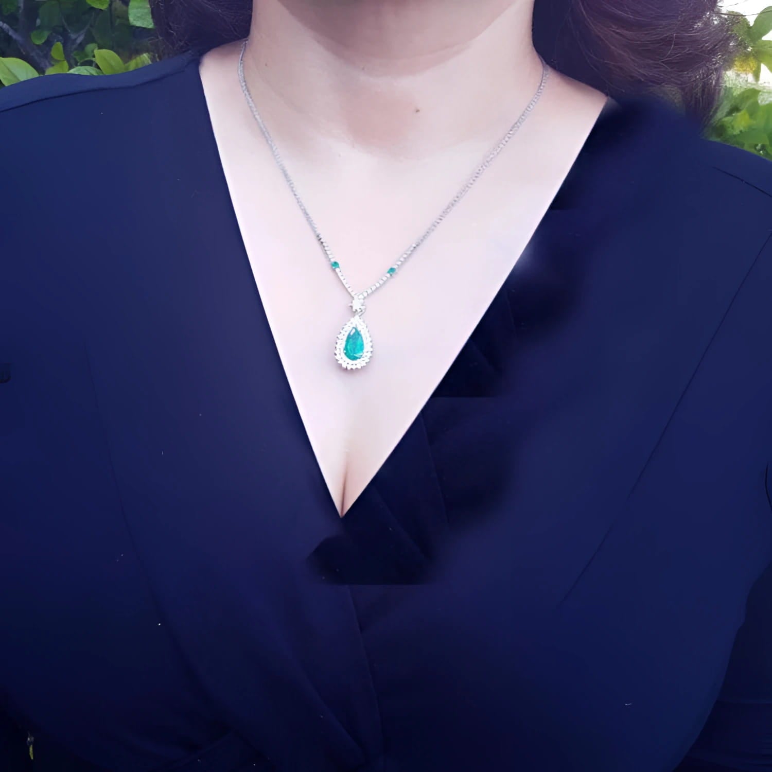 Pear cut emerald necklace with double diamond halo on 18K white gold chain, worn on a woman.