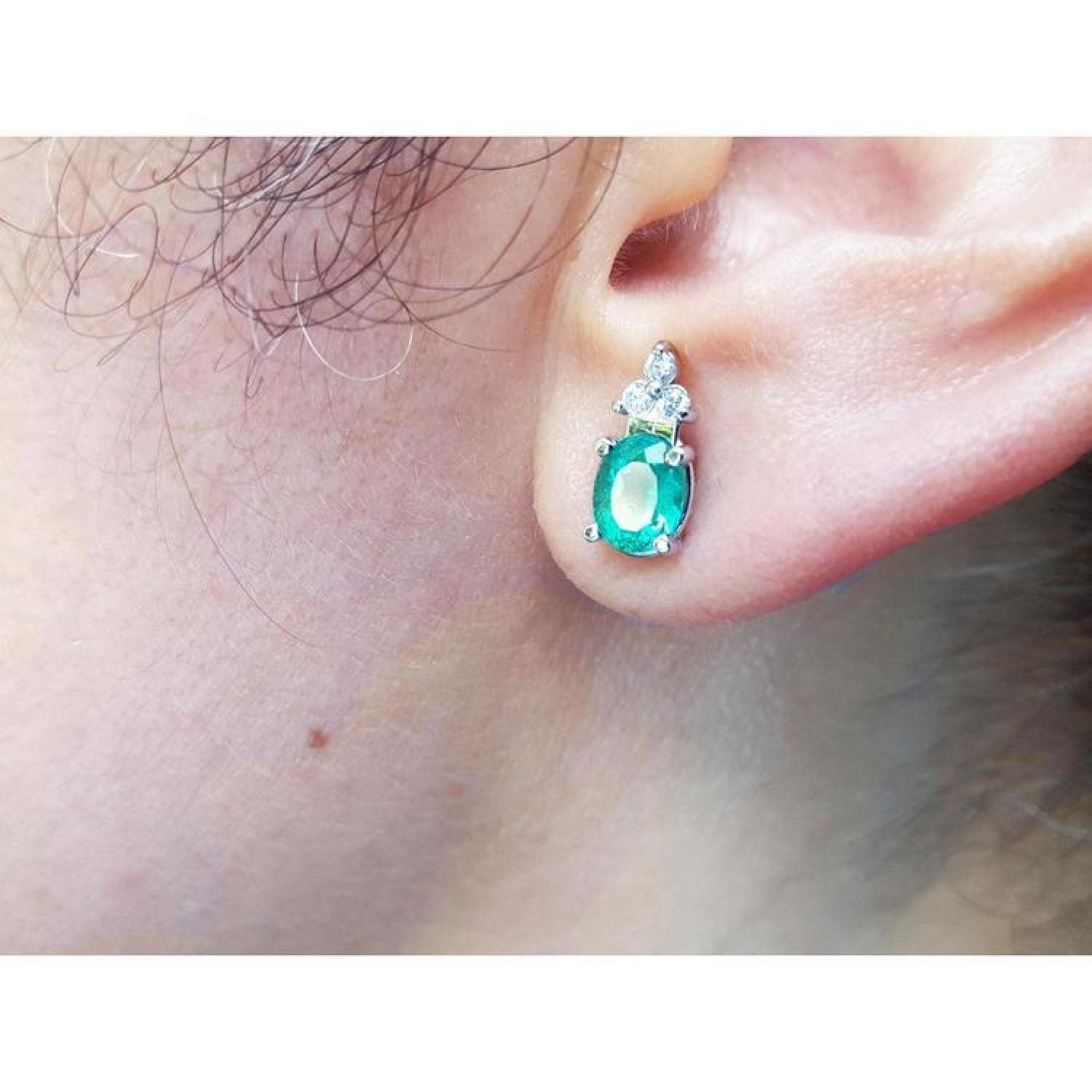 Oval natural emerald and diamond earrings in 14K white gold on an ear.