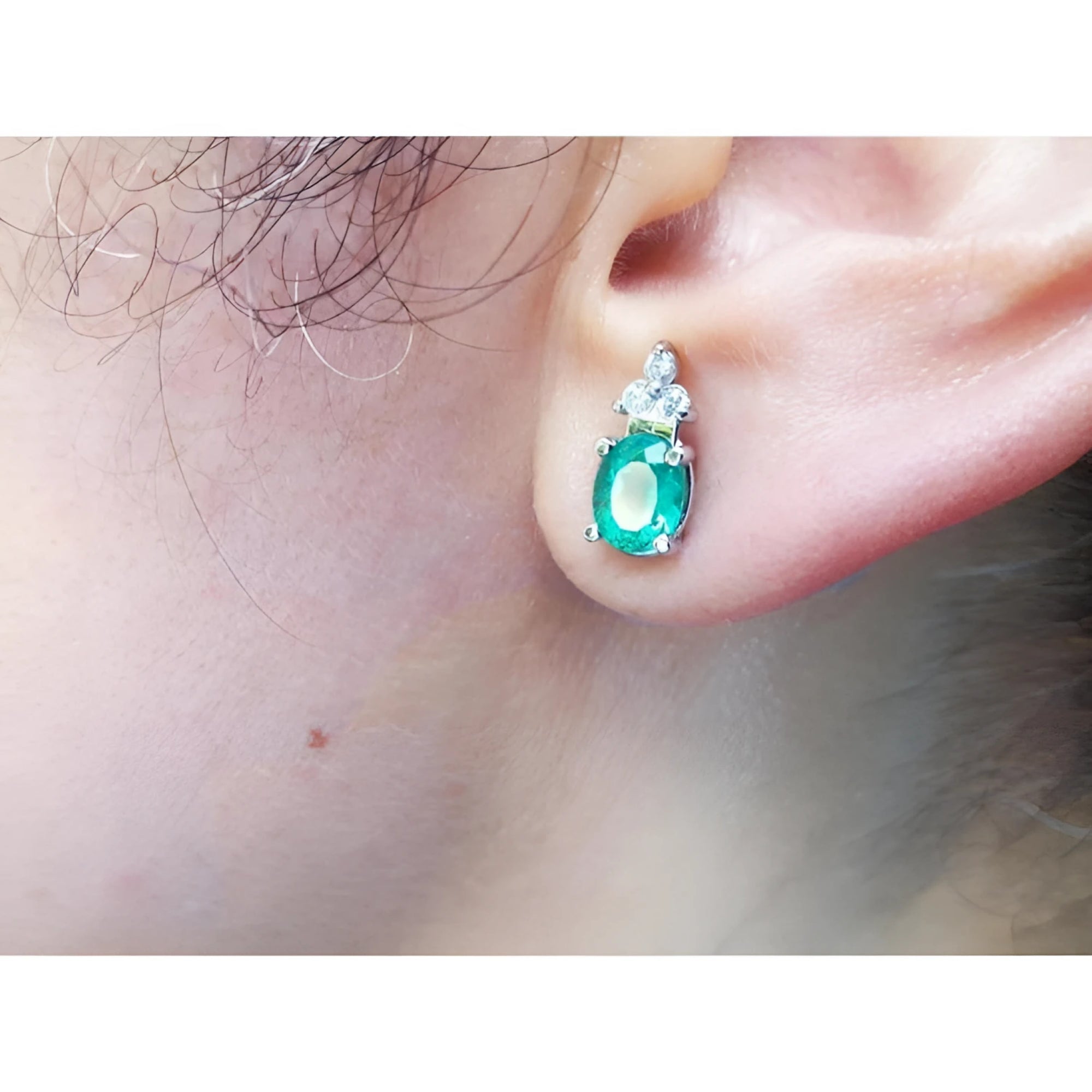 Oval natural emerald and diamond earrings in 14K white gold on an ear.