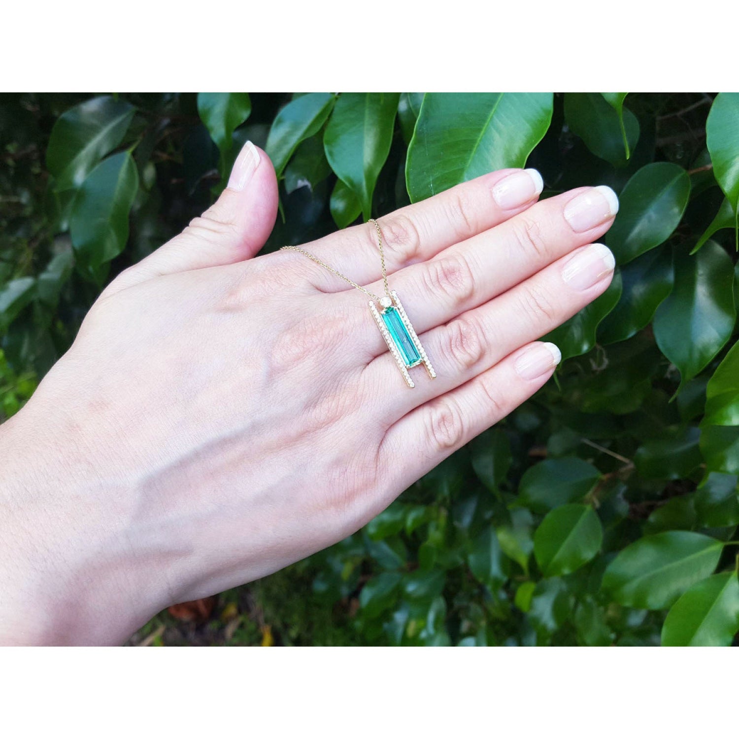 Emerald baguette cut necklace with natural diamonds and 14K yellow gold sliding tube bail shown worn on a person's hand, with a background of green leaves.