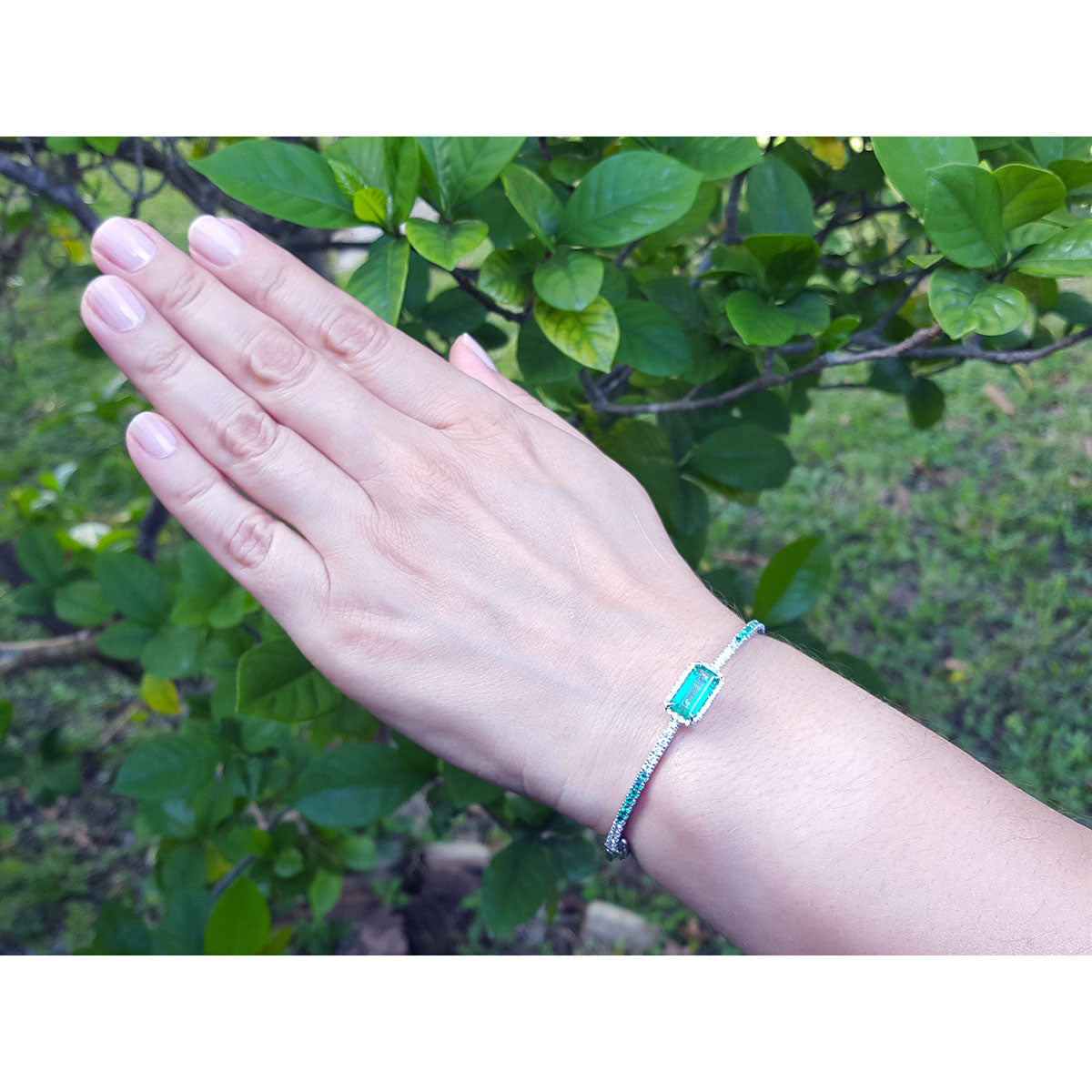 Hand wearing a natural emerald and diamond bangle bracelet with a green leafy background.