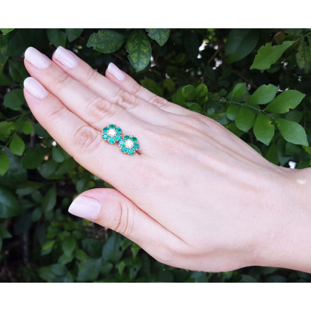 Hand displaying a pair of emerald and diamond cluster earrings with a leafy background.