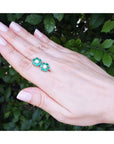 Hand displaying a pair of emerald and diamond cluster earrings with a leafy background.