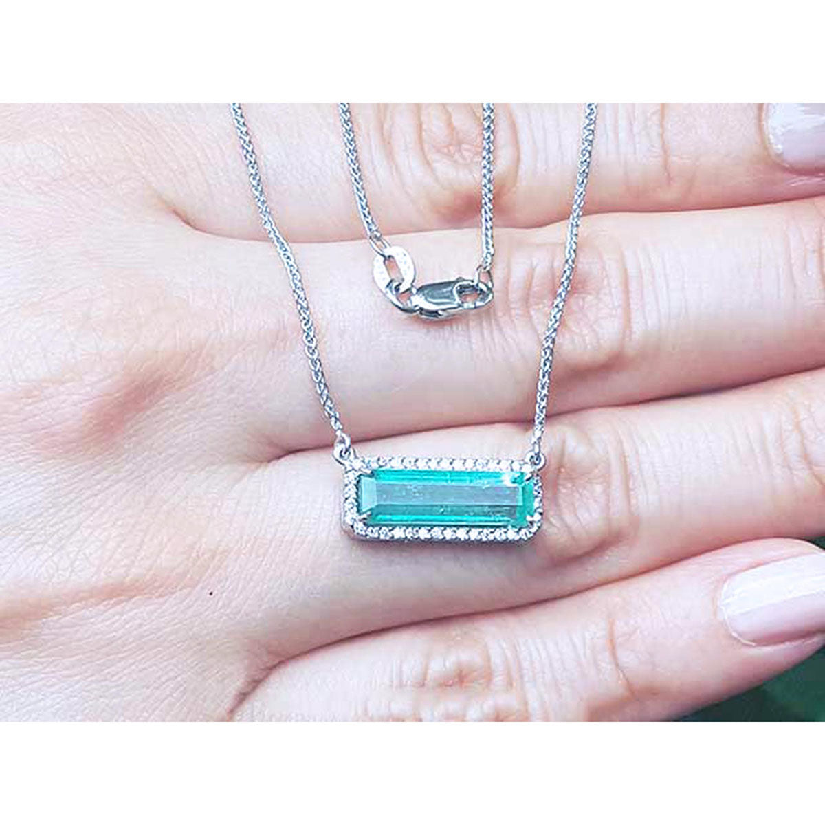 woman&#39;s hand showing a halo diamond Emerald necklace elongated east west orientation in 18k white gold