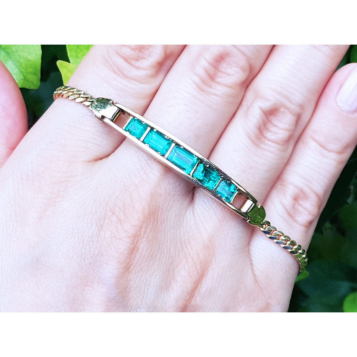 Gold bracelet with five natural colombian emeralds stones on a hand against a green leafy background.