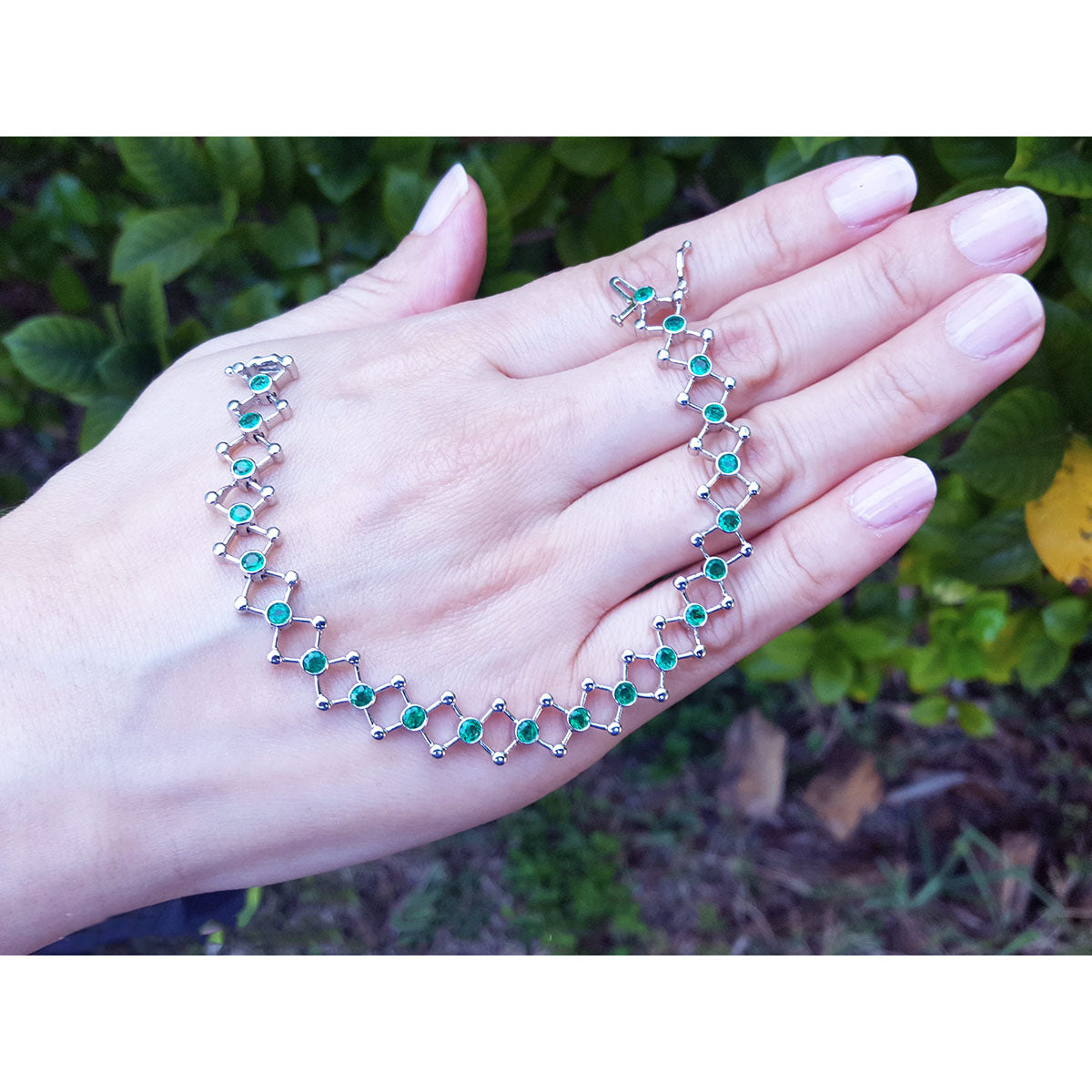 Hand displaying a white gold and emerald molecule-shaped bracelet with a blurred green outdoor background.