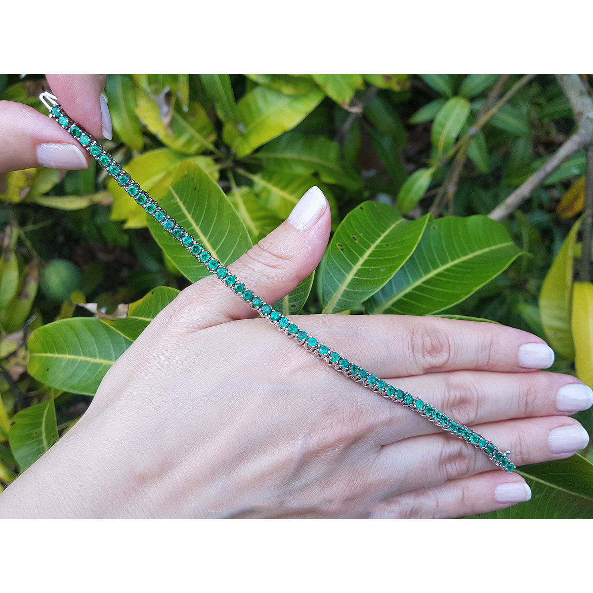 Emerald tennis bracelet held by a hand with green leaves in the background.