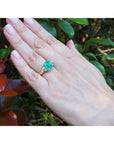 Hand wearing a Muzo Colombian emerald ring against a blurred natural background.
