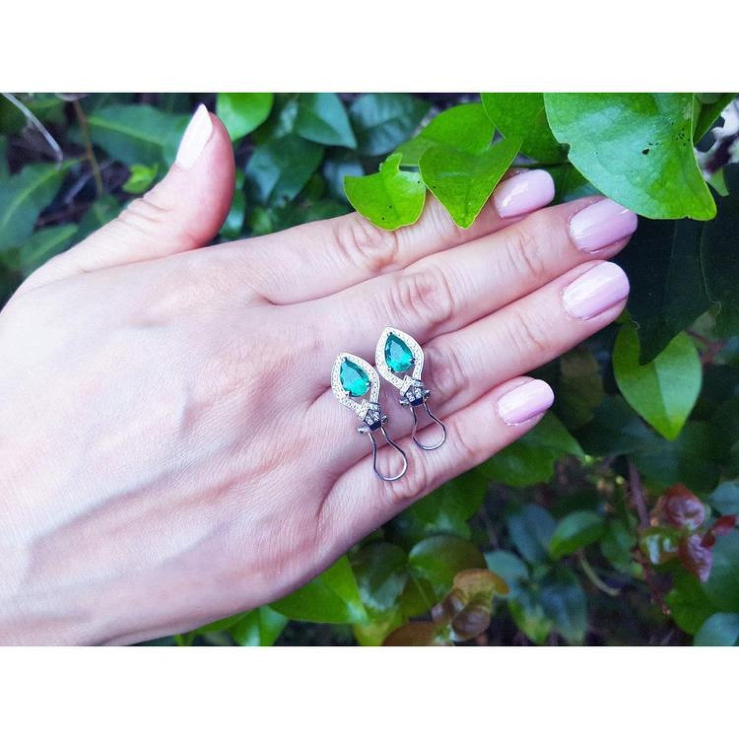 14K gold clip/post earrings with vibrant pear-shaped Colombian emeralds and diamond accents on a hand.