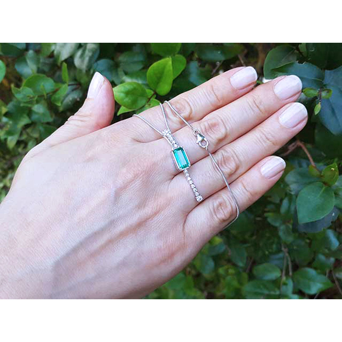 Hand displaying a natural emerald necklace with white gold chain against a green leafy background