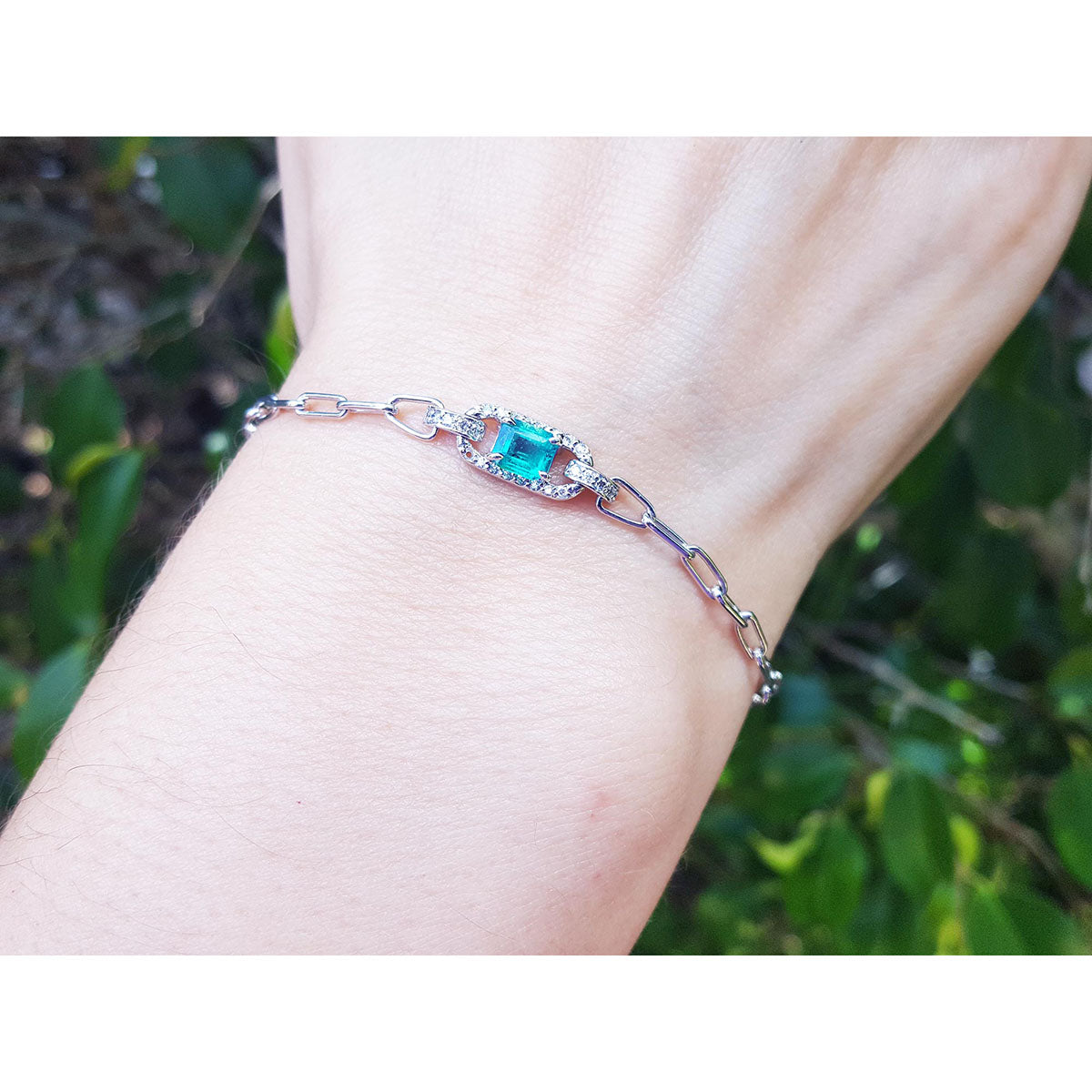 A paperclip bracelet with a natural colombian emerald on a wrist against a green leafy background.