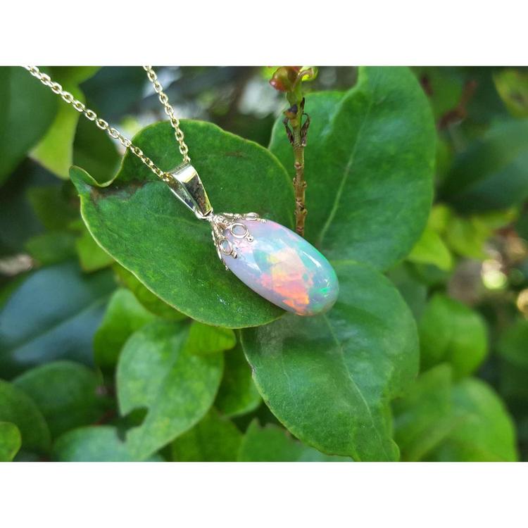 Ethiopian Welo opal necklace pendant with red and rainbow fire in 14k yellow gold, resting on green leaves.