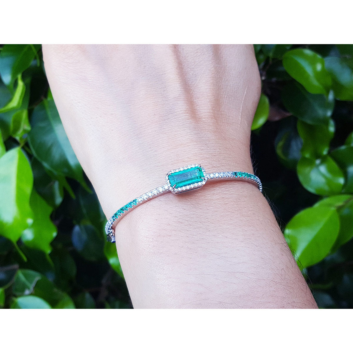 White gold bangle bracelet with a rectangular emerald on a wrist against a green leafy background.