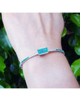 White gold bangle bracelet with a rectangular emerald on a wrist against a green leafy background.