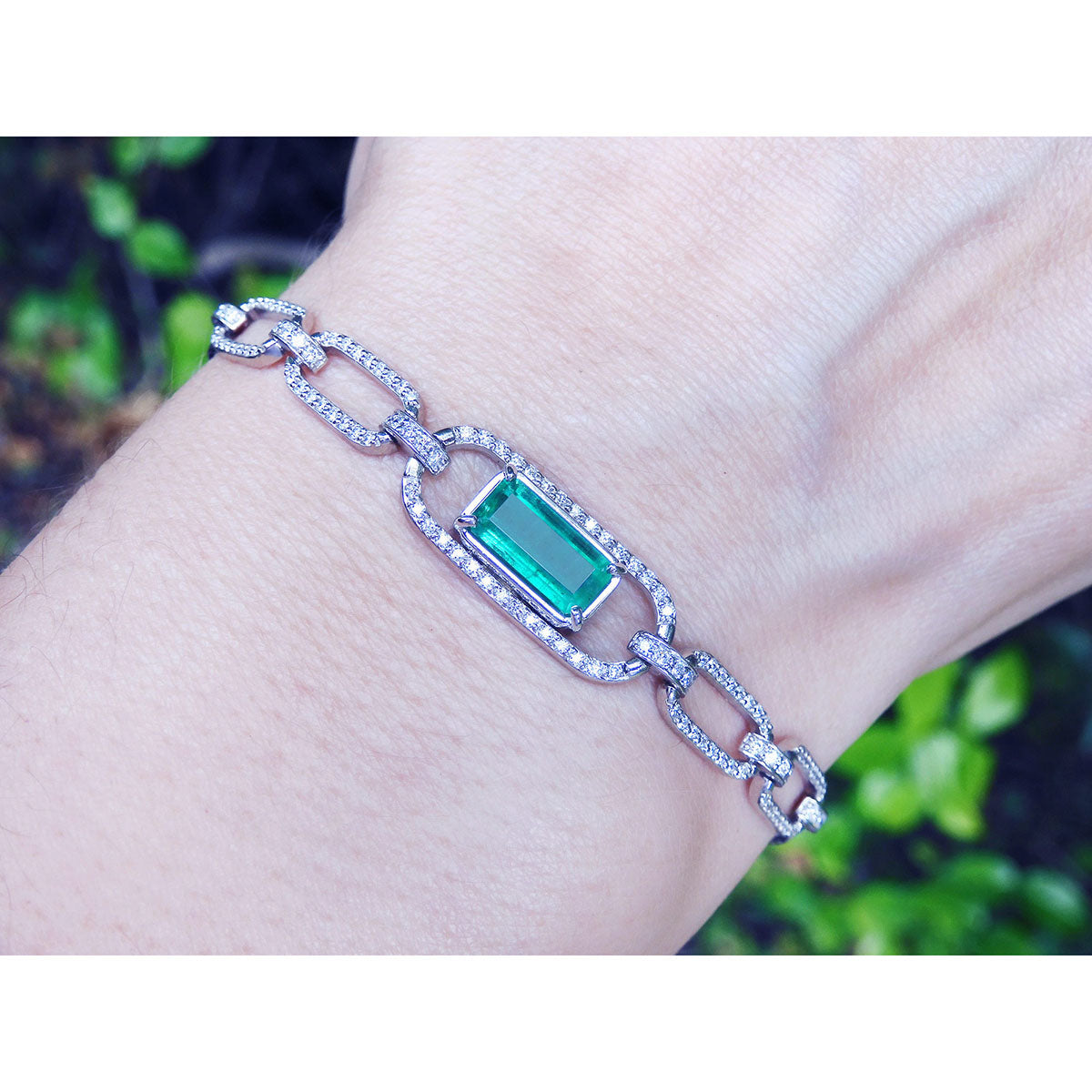 A white and yellow gold bracelet with a Muzo emerald gemstone on a wrist against a blurred natural background.