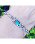 A white and yellow gold bracelet with a Muzo emerald gemstone on a wrist against a blurred natural background.