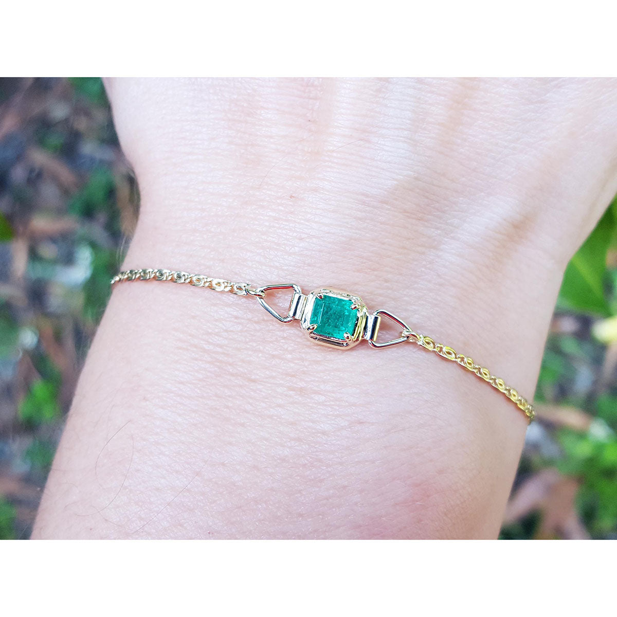 Gold bracelet with a square emerald on a wrist against a blurred natural background.