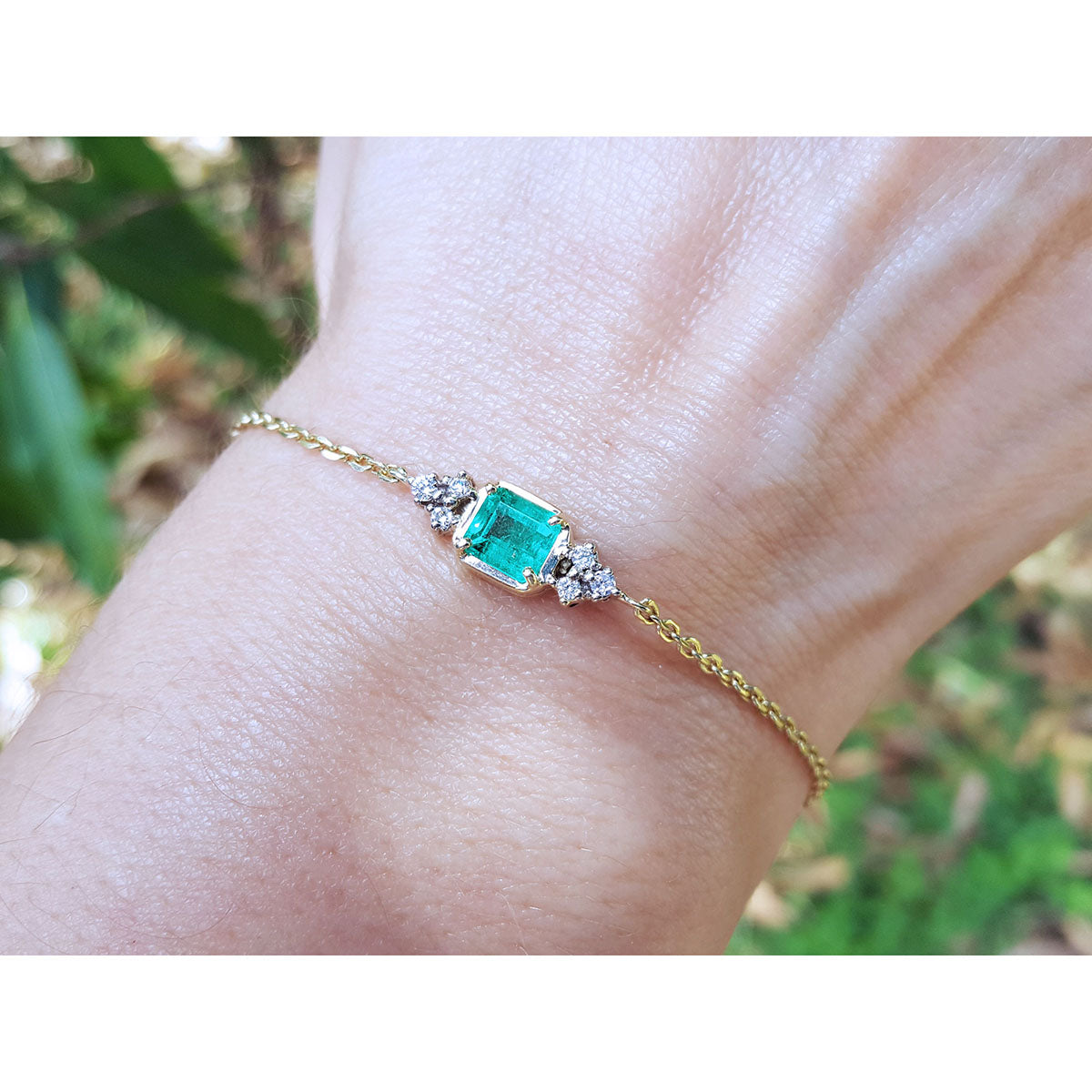 Gold bracelet with a emerald-cut emerald on a wrist against a blurred natural background.