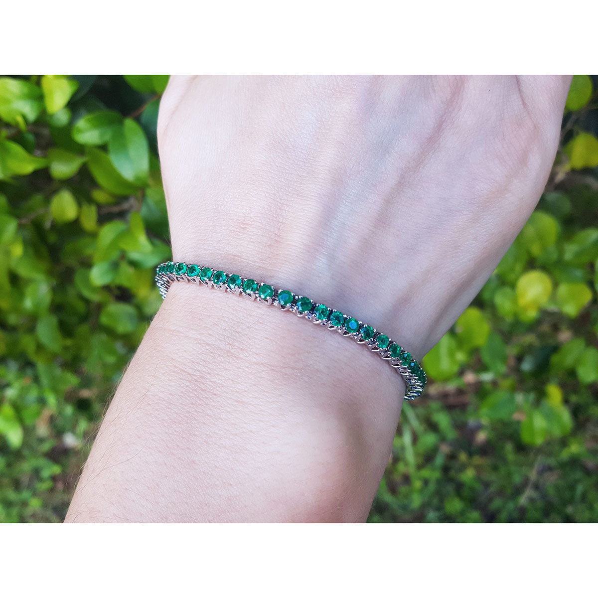 A round cut genuine colombian emerald tennis bracelet on a wrist with a blurred green leafy background