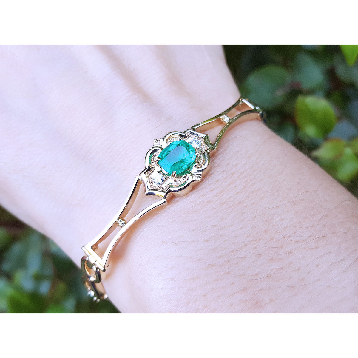 Gold bracelet with an oval cut natural emerald on a wrist against a blurred green background.