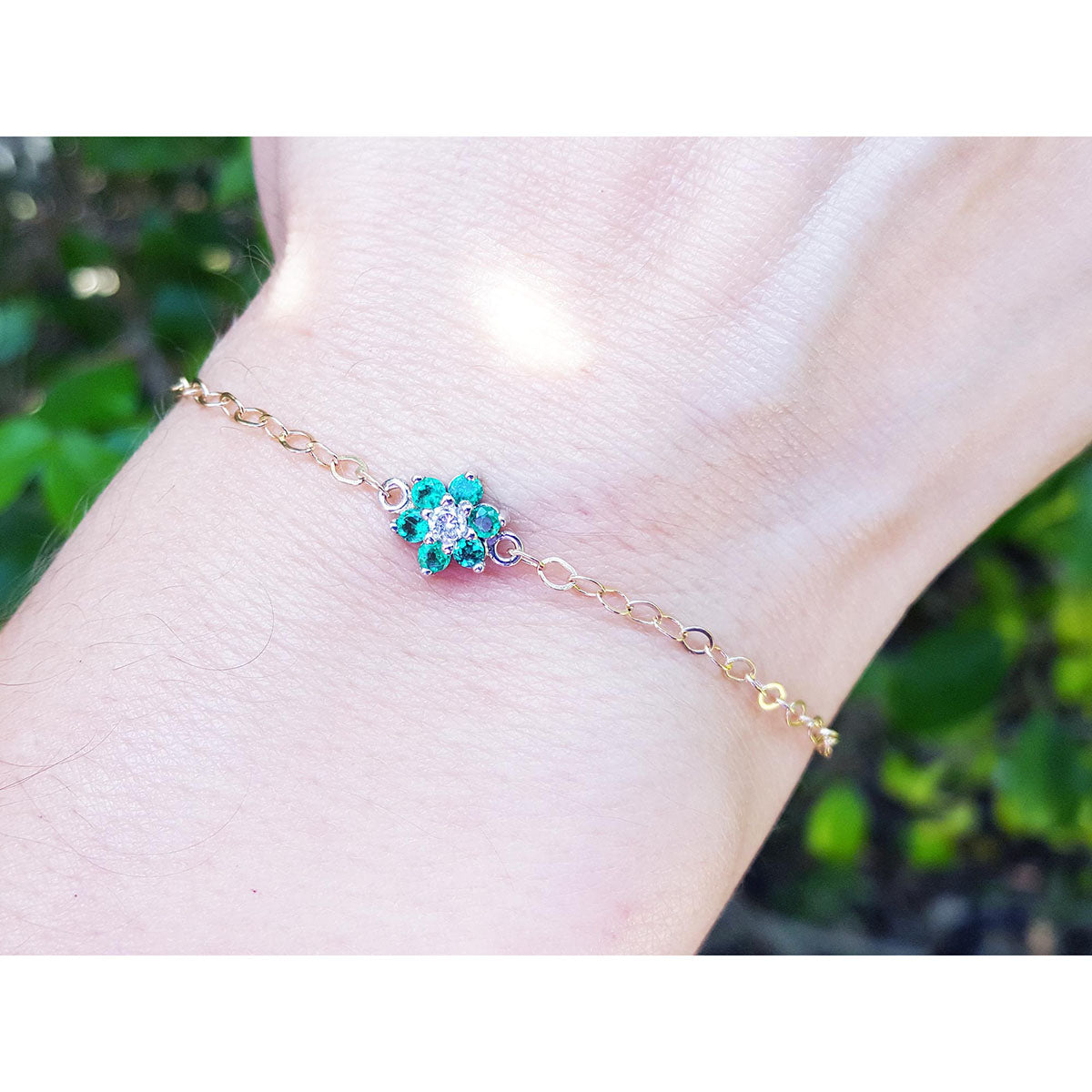 Gold bracelet with a emerald cluster flower charm on a wrist against a blurred green background.