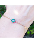 Gold bracelet with a emerald cluster flower charm on a wrist against a blurred green background.