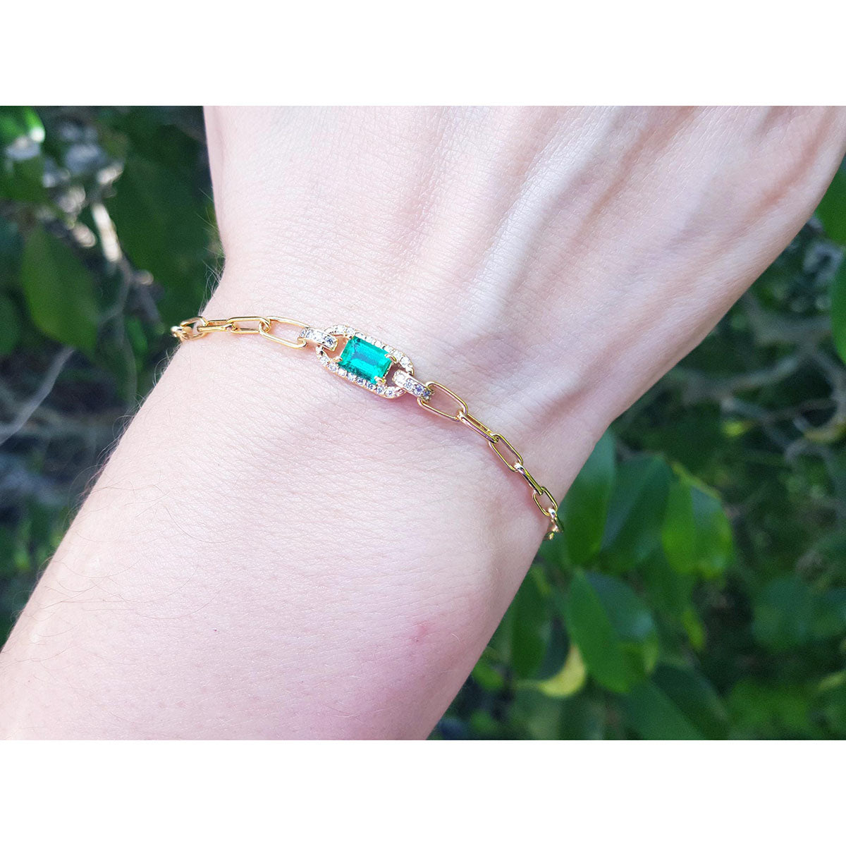 Gold bracelet with a rectangular emerald on a wrist against a blurred green background.