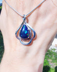 Silver pendant with a blue Welo teardrop black opal held on a person's wrist outdoors.