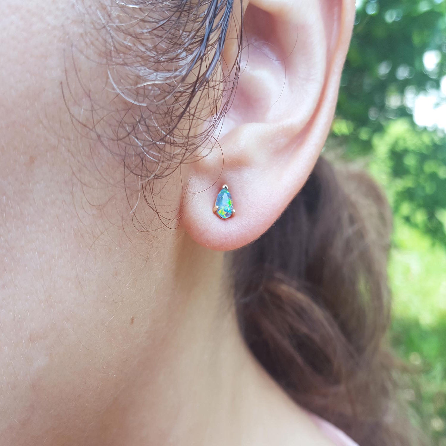 Close-up of an ear wearing a colorful teardrop opal earring with a blurred green background.