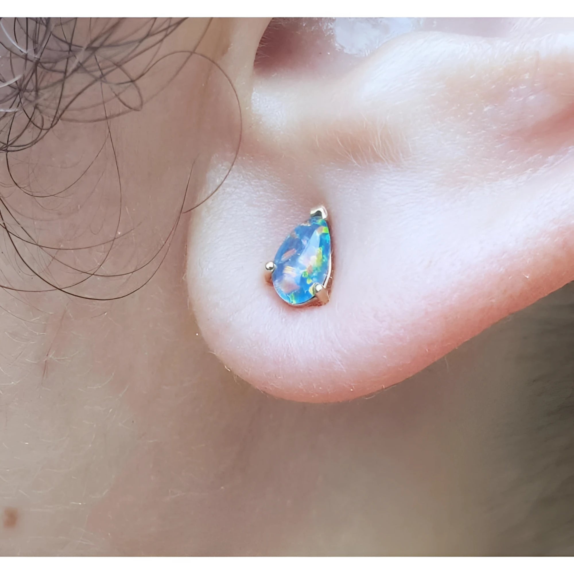 Pear-shaped opal doublet stud earring with red and blue play of colors, set in 14K yellow gold on an earlobe.