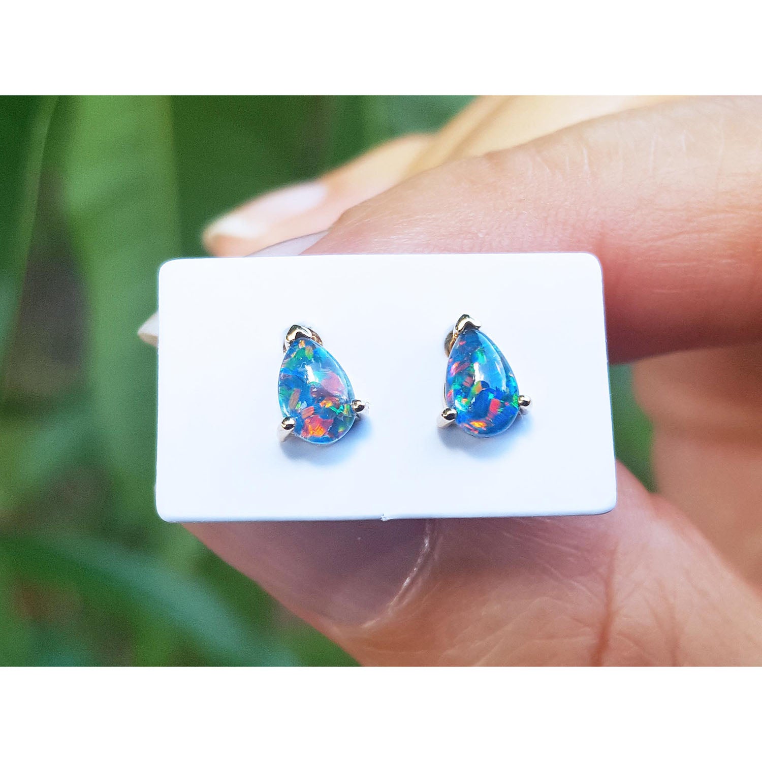 Teardrop-shaped opal earrings on a white card held by a hand with a blurred green background.