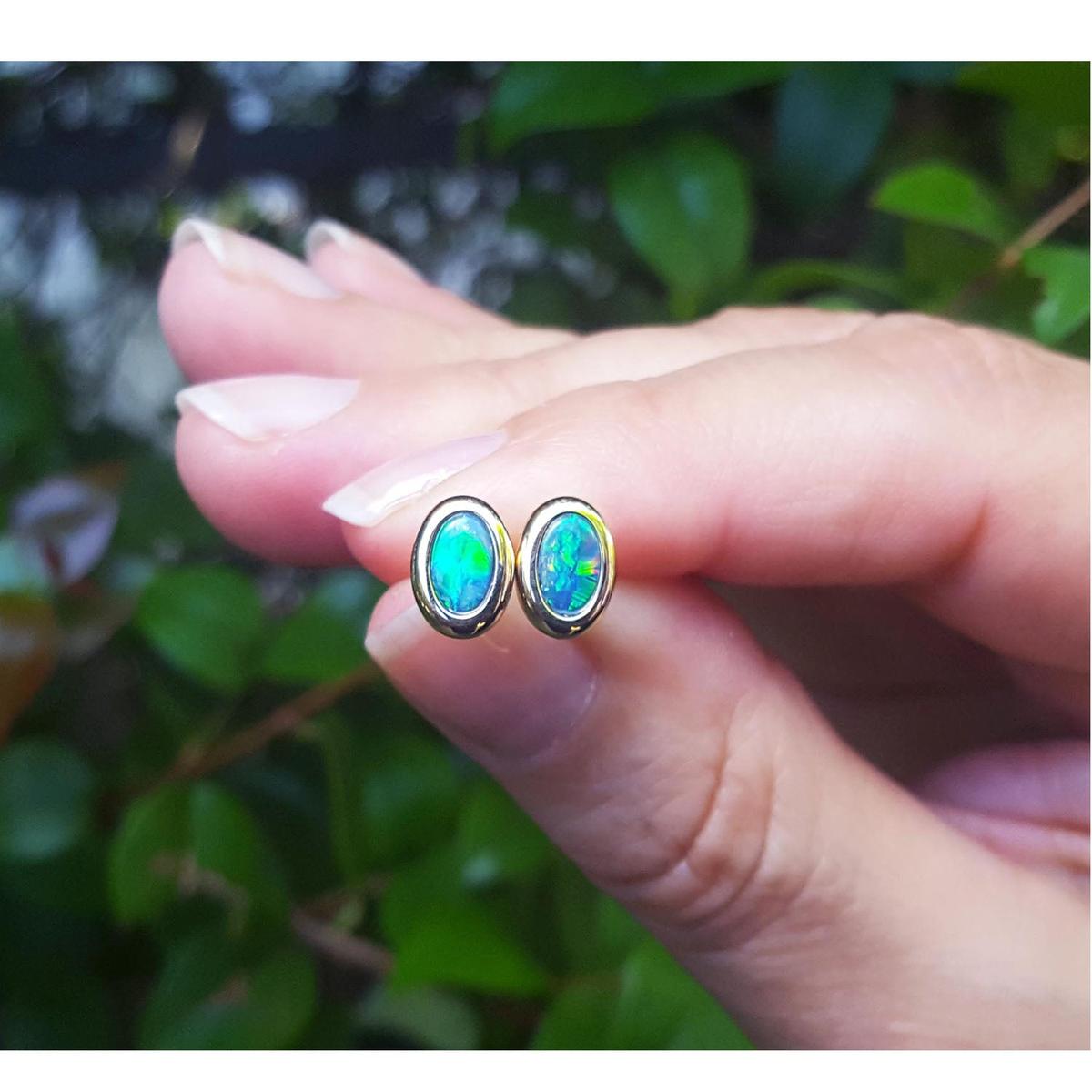 Opal stud earrings held between fingers with a blurred green leafy background