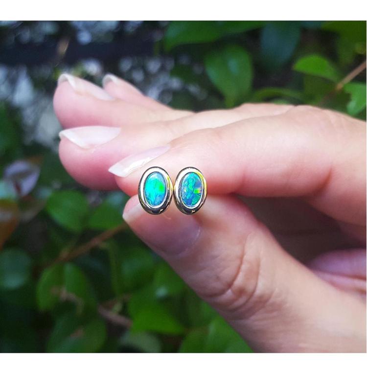 Opal stud earrings held between fingers with a blurred green leafy background