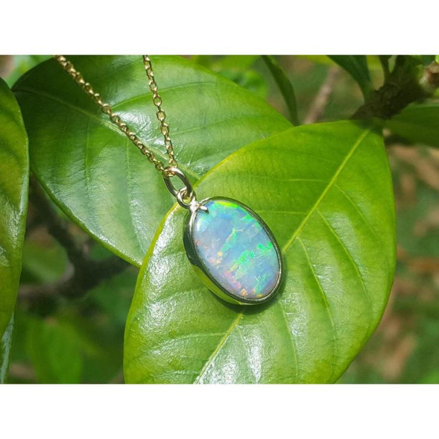 Oval Australian opal pendant with green-bluish rainbow play, set in 14K yellow gold, resting on a green leaf.