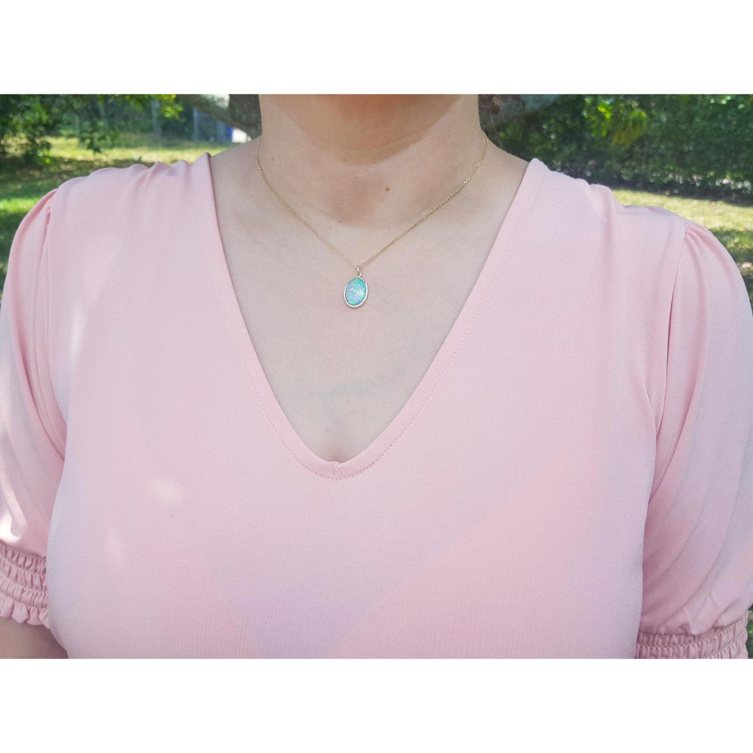 A woman wearing a pink top with a necklace featuring a blue opal pendant outdoors.