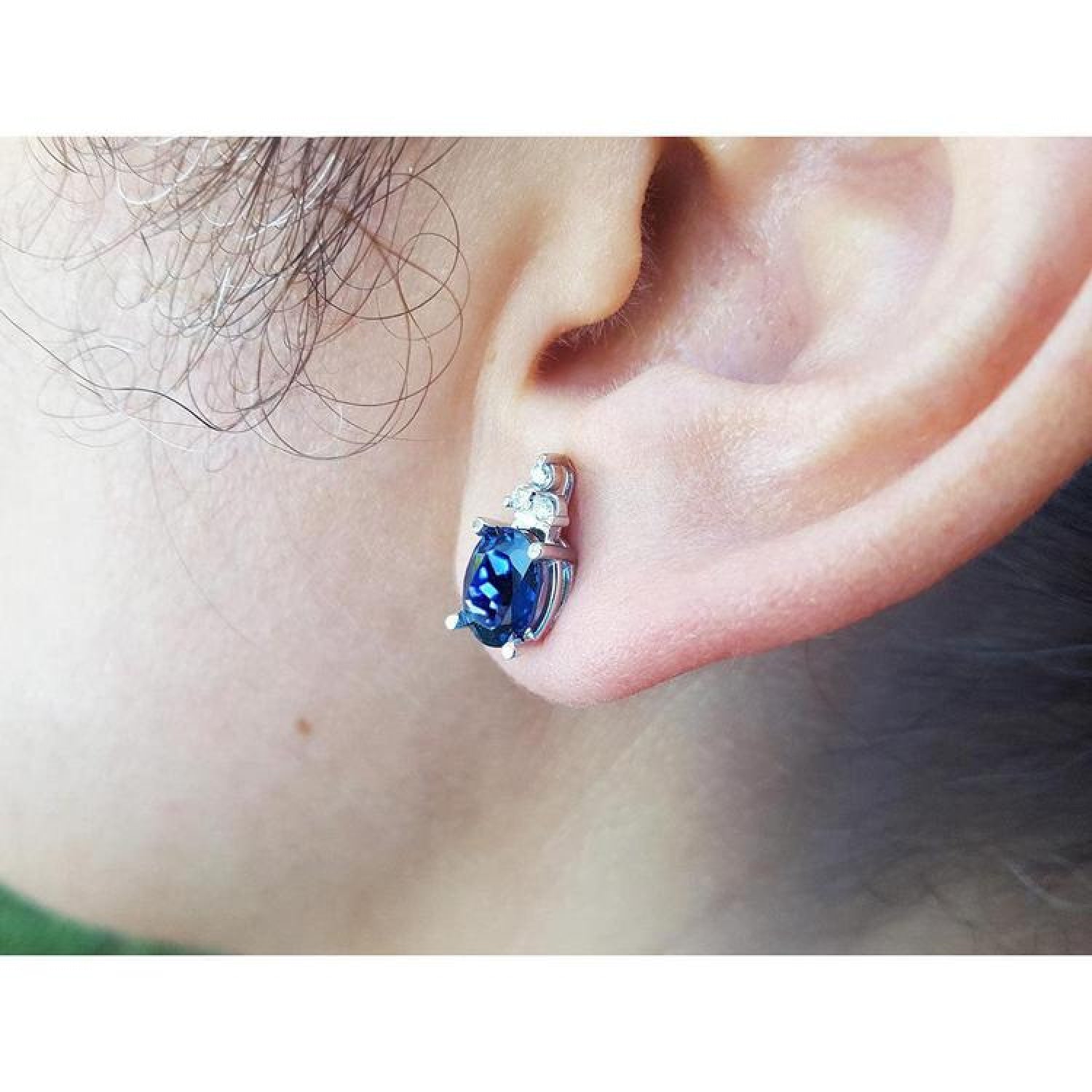 Oval blue sapphire stud earring with three small diamonds in 14K white gold on an ear.